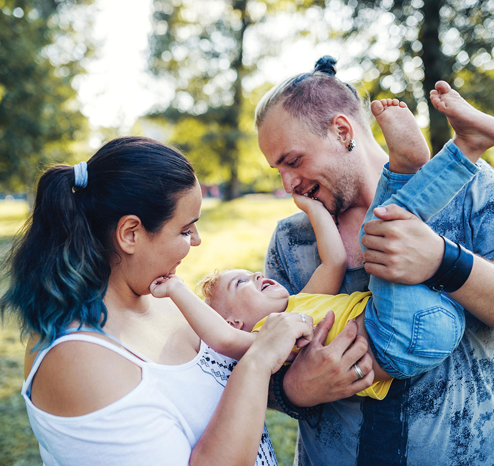 A couple joyfully holds their child in a sunny park filled with flowers